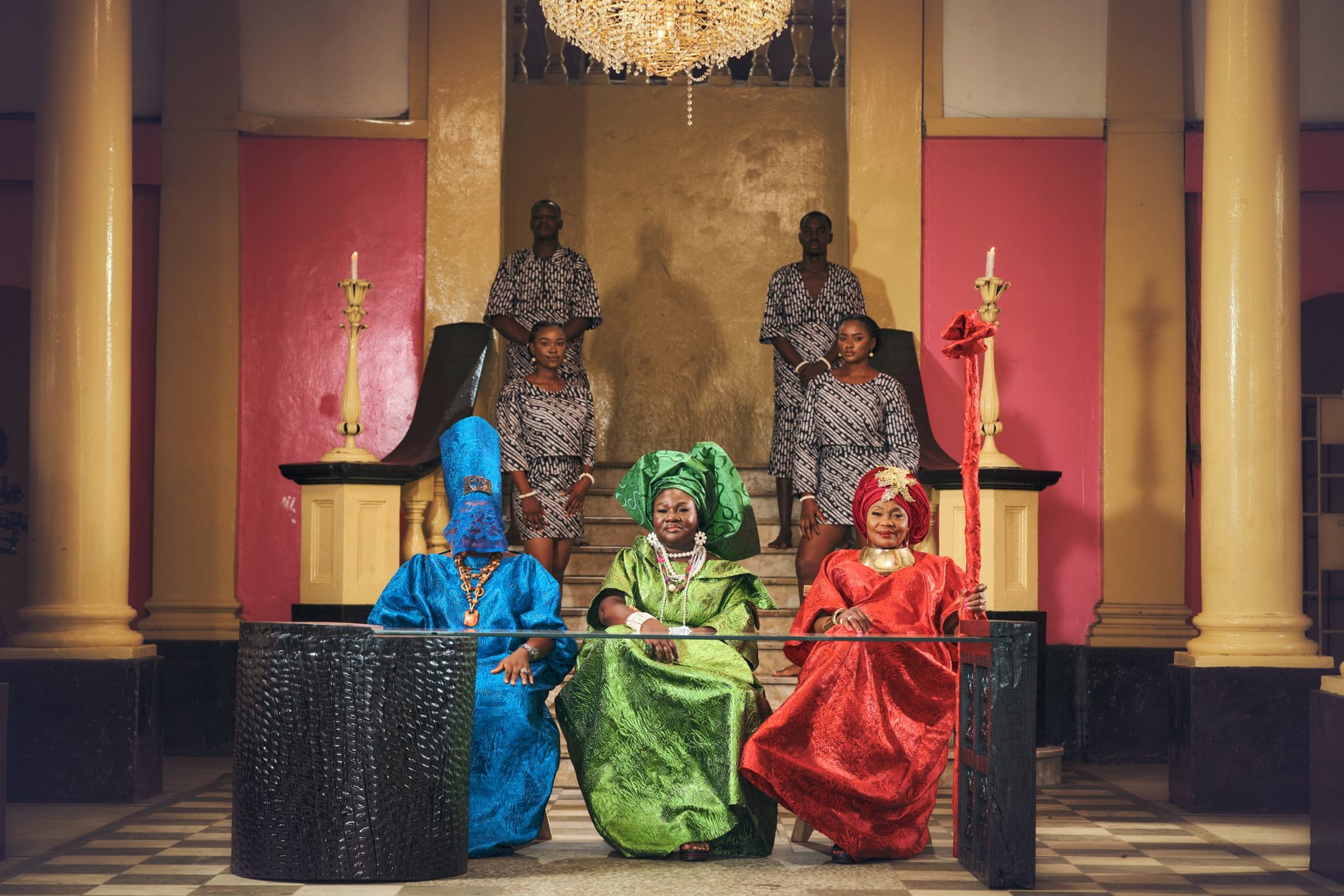 Three women in blue, green and red seated at the glass-topped table, with four attendants standing on the stairs behind them